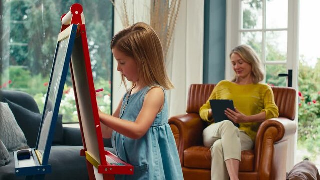 Family with daughter drawing picture on easel as mother with digital tablet watches from sofa at home -shot in slow motion 