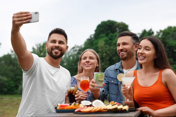 Happy friends with glasses of cocktails taking selfie at table outdoors