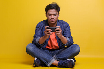 Focused Asian man in casual clothes playing games on smartphone, sitting crossed legs isolated over yellow background.  E sport. 