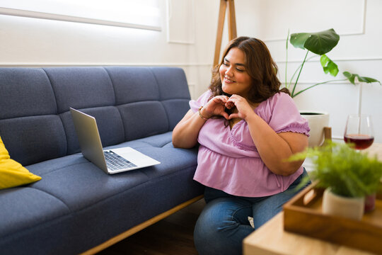Plus size woman making a heart shape with her hands during a videocall at home with her virtual date - Powered by Adobe