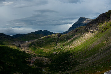 Fototapeta premium Sunlit green mountain slopes on the way up to Rabothytta, Okstindan, Hemnes, Helgeland, Northern Norway. Green mountains of Nordnorge. Sunny patches on dark mountains. Cloudy dramatic skies and light.