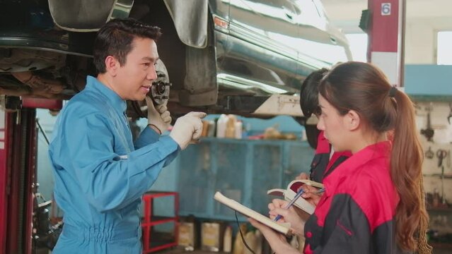 Asian male professional automotive engineer supervisor describes car wheel and suspension repair work with mechanic worker staffs team in fix service garage, specialist occupations in auto industry.