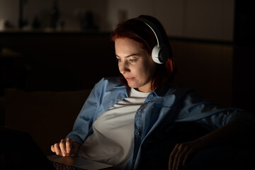 Focused programmer woman in dark room sits on chair work on laptop . Workaholic freelancer female sitting comfortably with computer on laps, wear headphones working on graphic design for app at night