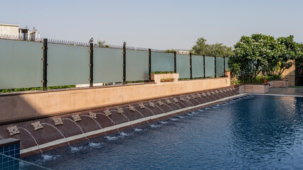 A fragment of a swimming pool with blue water. Water jets are pouring the perimeter. The fence is made of frosted glass. Green trees nearby.