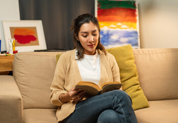 Asian beautiful woman sitting reading book on cozey couch sofa in living room with warm light at night. People female stay home part time relax at home.