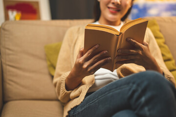 Fototapeta premium Close up female hands holding book turning the page. Young woman sitting on cozey couch sofa reading book in living room with warm light at night. People female stay home part time relax at home.