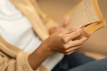 Close up female hands holding book turning the page. Young woman sitting on cozey couch sofa...