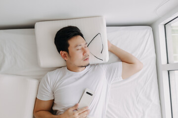 Asian man falling asleep with smartphone in hand on white bed.
