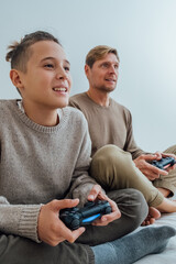 Mature father with teenager boy playing video games on console use joystick controller sitting on sofa enjoy game smile laughing.