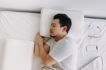 Asian man sleep lie down on white bed at room condo with eyeglasses and smartphone.