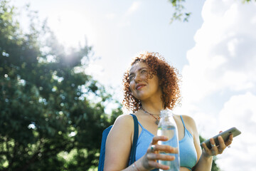 Low angle view of female in blue sport top on her way to gym for morning pilates training and yoga practice, holding smartphone and bottle of fresh water pictured on background of sunny and cloudy sky