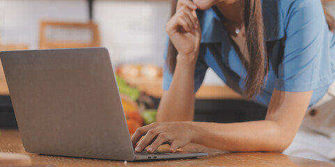 Fototapeta premium A young woman with a beautiful face in a blue shirt with long hair eating fruit sitting inside the kitchen at home with a laptop and notebook for relaxation, Concept Vacation.