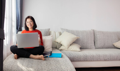 In the morning, the Asian elderly woman is sitting on the sofa and using the computer.