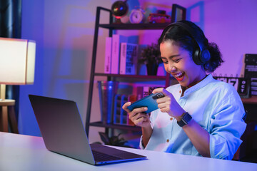 Excited Young Asian woman wearing headset and playing online game on smartphone at night modern...