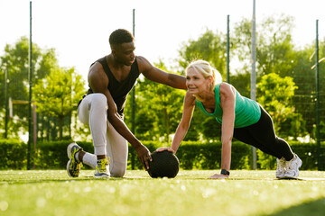 Young trainer assisting mature woman while she doing push ups during workout in park - Powered by Adobe