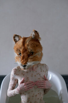 Young woman with red fox head sitting in bathtub filled with water and covering her breast with hands as if protecting 
