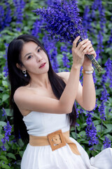 Asian woman in white dress sitting amidst in flowers field. Beautiful girl holding bouquet flowers in hands. Happy woman enjoying in purple flower field and nature sea of fog. Embrace nature,wind.
