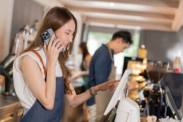 Asian coffee shop female employee accepts a pre-order on a mobile phone call while using a tablet computer in a cafe. Woman waiter talking on the cellphone. Home food delivery concept