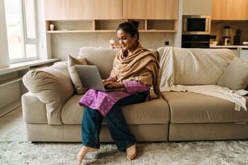 Obraz premium Young indian woman smiling and using laptop while sitting on couch