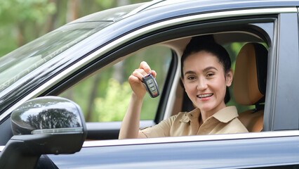 Black-haired businesswoman boasts showing keys from recently purchased car. Woman sitting in cabin holds keys with delighted and amused expression.