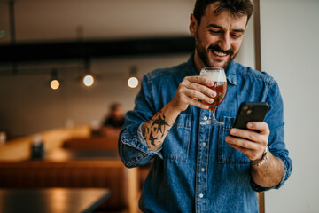A man in a denim shirt standing by the window indoors, drinking a beer and using a phone