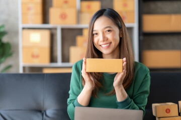 Smiling young Asian business owner woman prepare parcel box and check online orders of product for deliver to customer on laptop computer at home. Shopping Online concept. Startup small business SME.