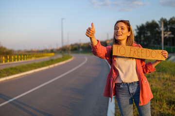 Woman is hitchhiking on roadside trying to stop car. She is holding cardboard with inscription.