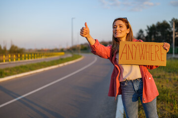 Woman is hitchhiking on roadside trying to stop car. She is holding cardboard with inscription.