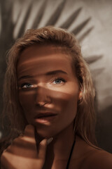 Close-up portrait of a stunning blonde in a black bikini, with palm branch shadows and sun-kissed skin, evoking a tropical vibe