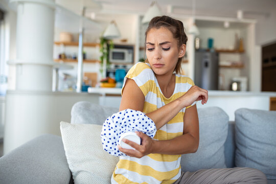 Young woman is sitting at home on her sofa and touching the her elbow while suffering from elbow pain, placing ice pack on hurt spot