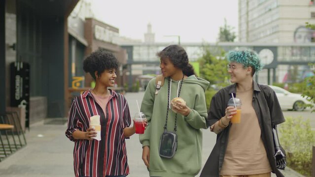 Medium shot of three diverse teen girls walking on city street towards camera, having takeout food and drinks, smiling and chatting while spending summer day together outdoors