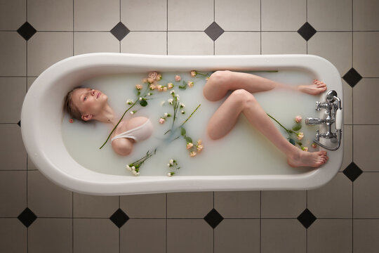 Top view of attractive young woman in swimsuit looking away pensively while relaxing in tub filled with milk and roses 