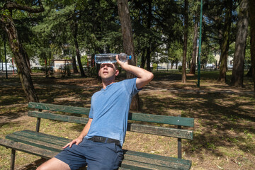 Man suffering from heat wave Charon in South Europe. Caucasian male presses bottle of water to face to cool off suffering from heat sitting outdoors - obrazy, fototapety, plakaty