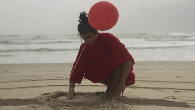 An adult girl in a red dress sitting on the sandy beach and playing with the sand forming a sand castle during the daytime