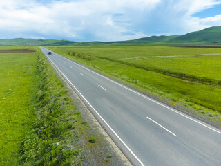 Fototapeta na wymiar Black passenger car is driving along an asphalt road along green fields, mountains on a sunny day. Photographing from a drone. Concept of travel