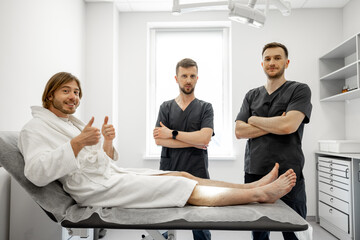 Two confident surgeons talk with a young male patient lying on a couch with trauma in medical office. Concept of traumatology and rehabilitation