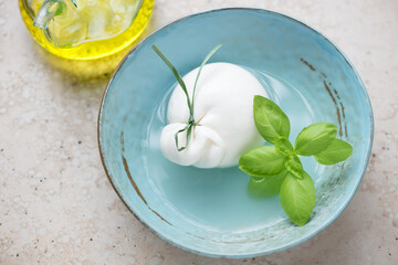 Burrata cheese and fresh green basil in a turquoise bowl, horizontal shot on a beige stone background