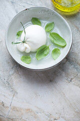 Grey bowl with burrata cheese in brine and green basil, vertical shot on a grey granite background with space, high angle view