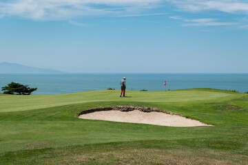 Campo de golfe com um golfista a jogar pr&oacute;ximo a um bunker de areia com o mar ao fundo num dia ensolarado