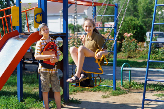 schoolchildren, a teenage boy and a girl play on the playground in the city park after school, a girl rides on a swing, a bright summer day