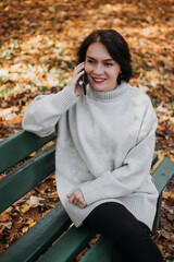 Smiling woman sitting on bench in autumn park using phone