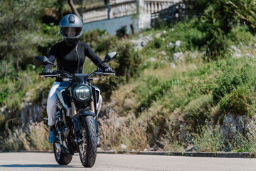 Fototapeta na wymiar Low angle of anonymous confident female motorcyclist in protective helmet and leather gloves riding modern motorbike on road near green trees in summer countryside