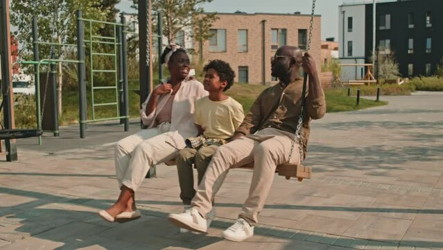 Stab shot of delighted modern African American family of three swinging on playground swings together while spending leisure time outside in summer