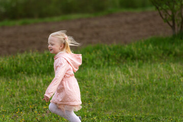 Happy running little European girl with blonde hair in pink outfit running across field or meadow. Spring time