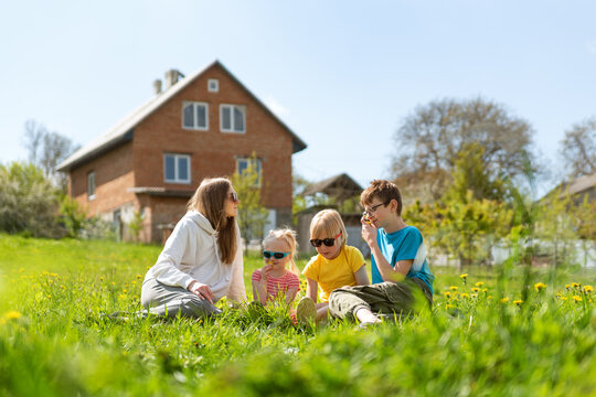 Mother and three children sitting on the grass in clearing with dandelions. Family picnic in backyard of house.
