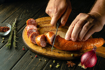 The chef cuts a meat veal sausage with a knife on a wooden cutting board. The concept of preparing delicious sandwiches for breakfast on the kitchen table