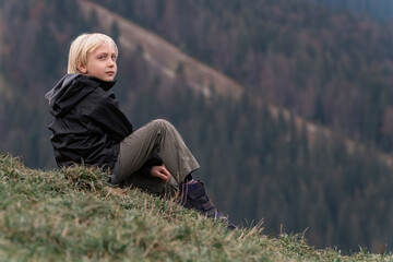Naklejka na ściany i meble Child in the mountains outside. Travel with children. Blond boy sits on hillside and looks into the camera