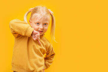 Portrait of blonde preschool girl with two tails in yellow sweatshirt on yellow background. Child points down.