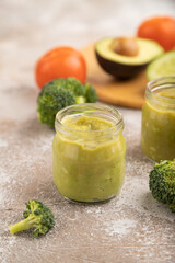 Baby puree with vegetable mix, broccoli, avocado in glass jar on brown concrete, side view, selective focus