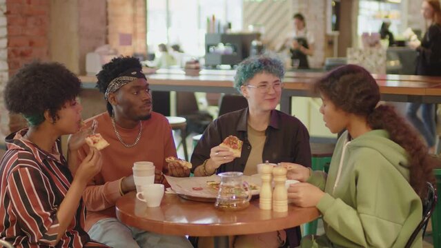 Company of multi-ethnic gen Z friends sitting together at cafe table, eating delicious pizza and chatting